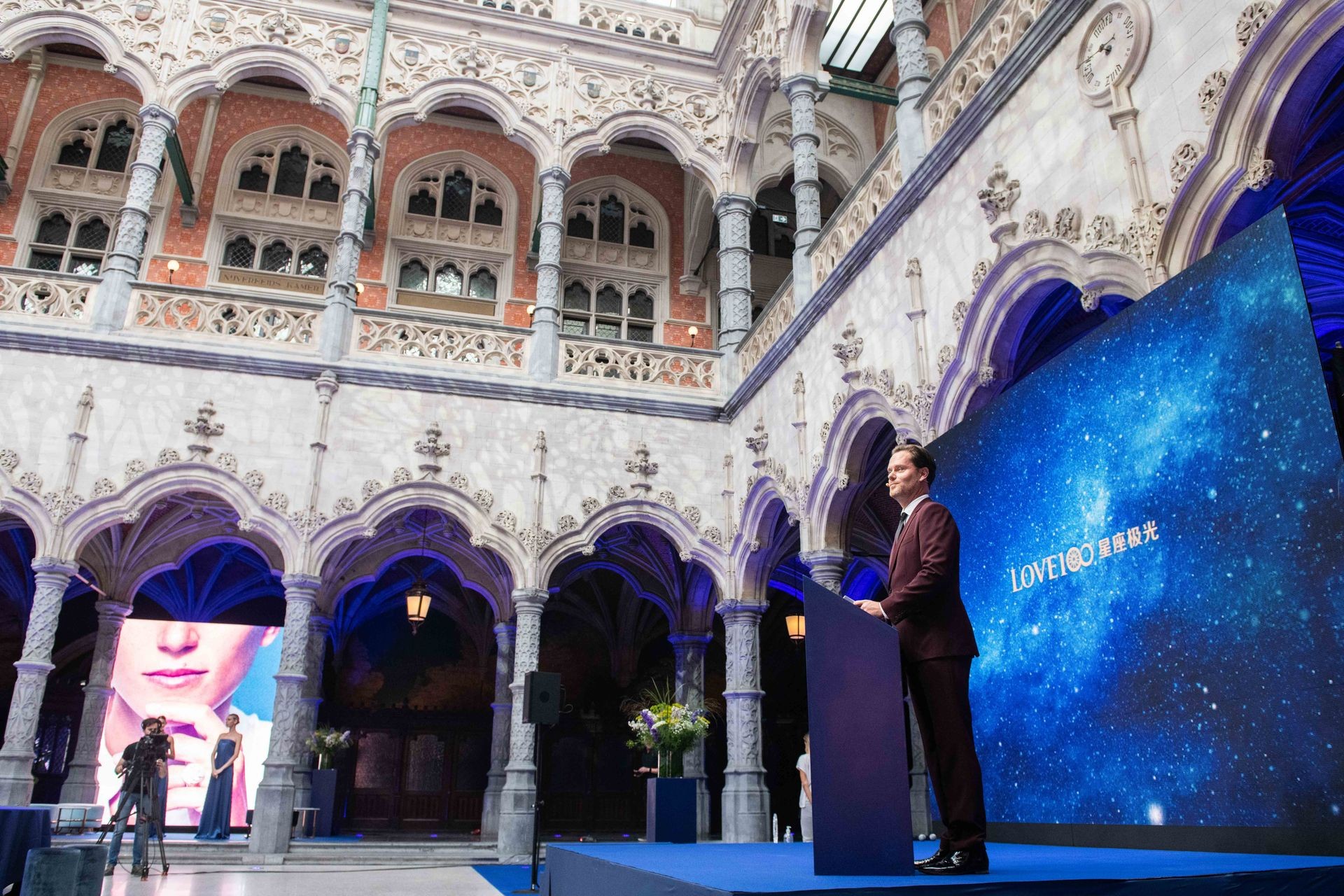Person speaking at a podium in an ornate, historic courtyard with a large blue display screen.